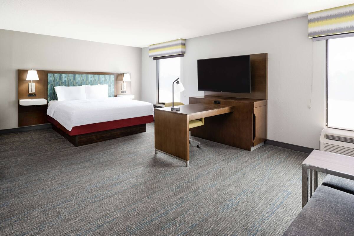 A hotel room with a bed, desk, TV, and a window with blinds.
