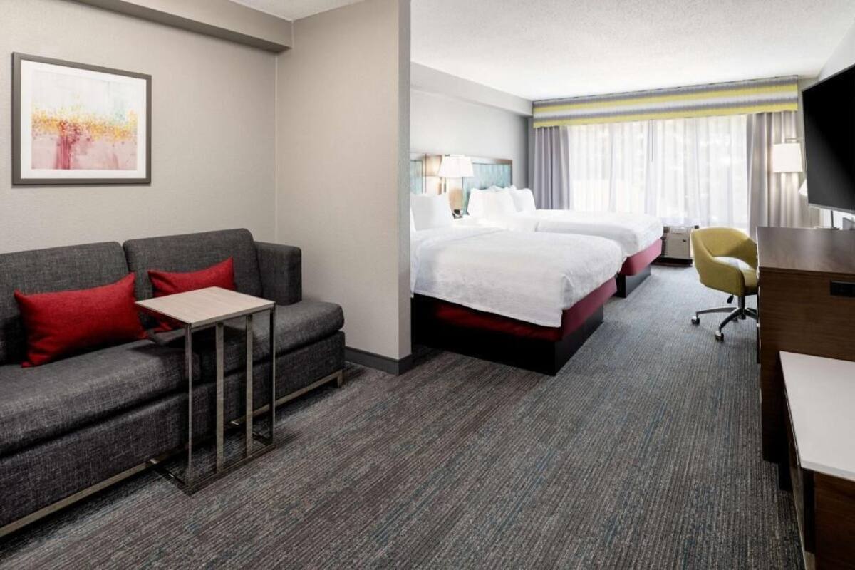 A hotel room with a grey sofa, a small table, a bed with white bedding, a TV, and a window with curtains.