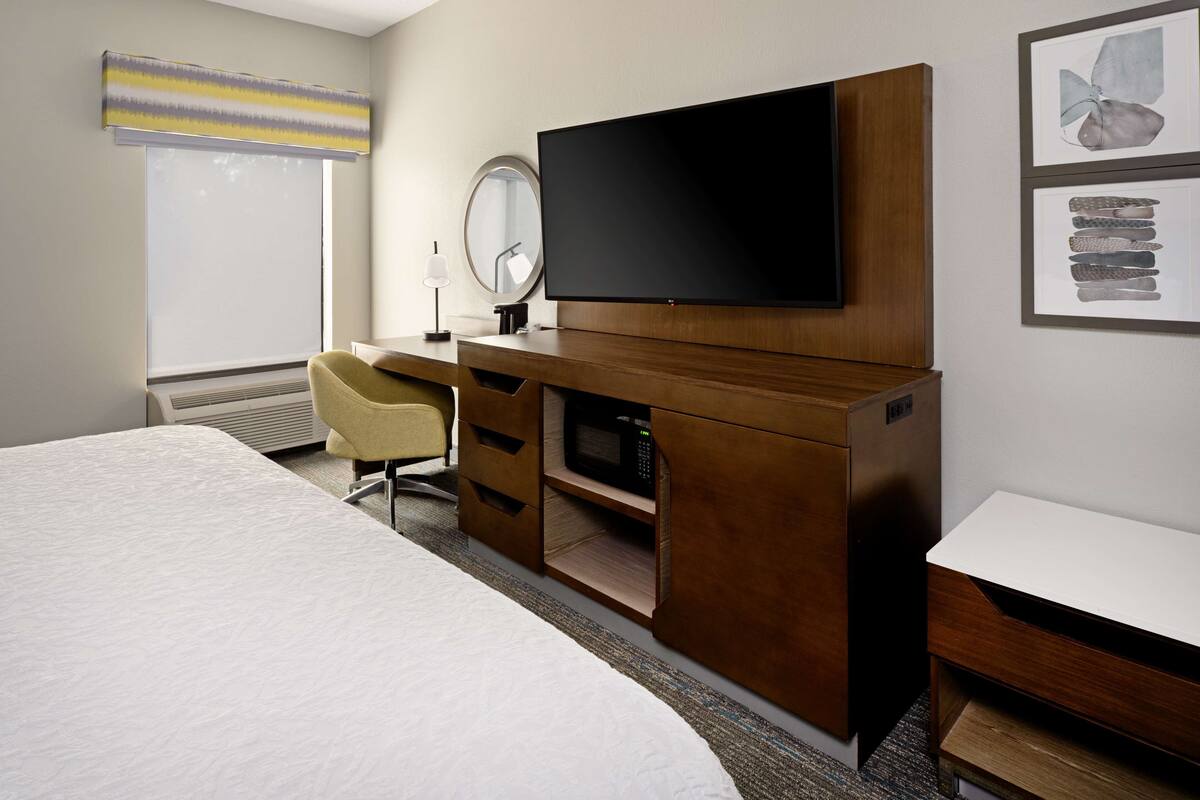 A hotel room with a bed, a desk with a flat-screen TV, a microwave, a chair, and a large window with blinds.