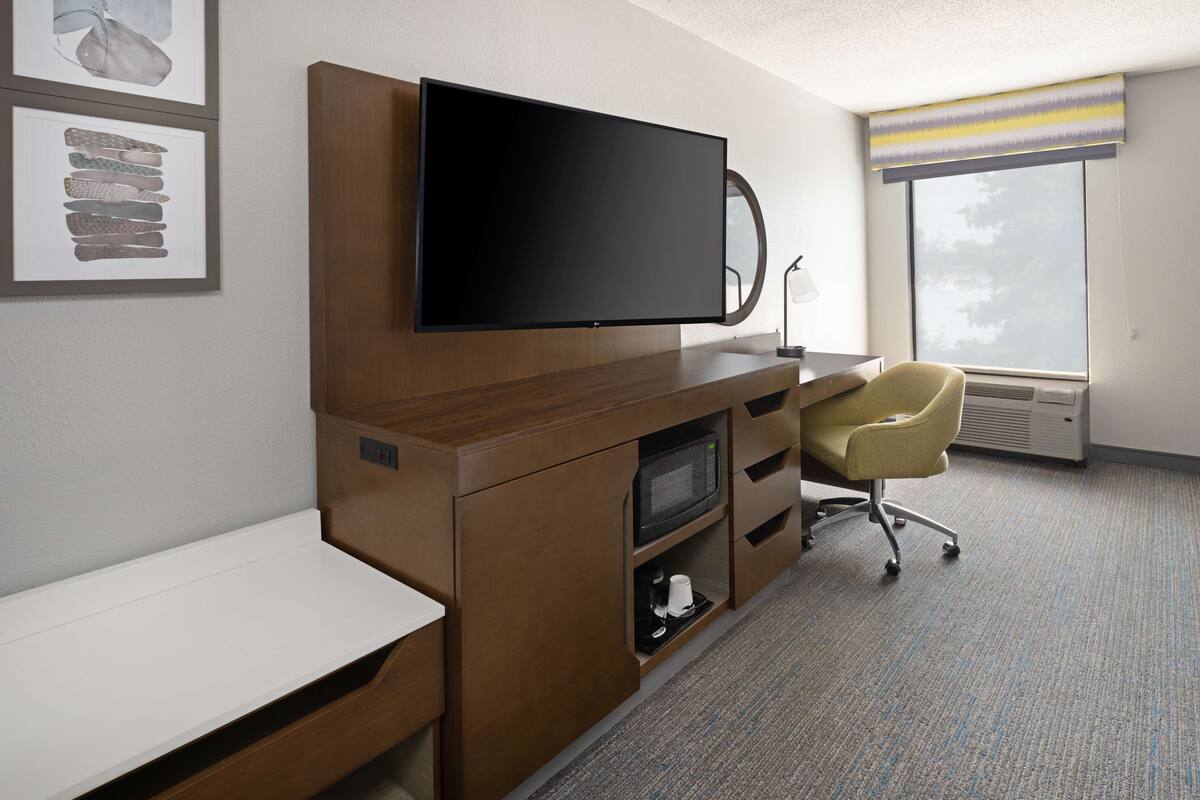 A hotel room with a flat-screen TV mounted on a wooden cabinet, a desk, a chair, and a large window with a view of trees.