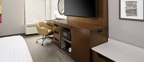 A hotel room with a bed, a desk with a flat-screen TV, a chair, a microwave, and a bench.