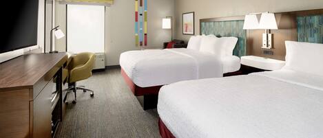 A hotel room with two beds, a desk, a chair, a TV, and a window with blinds.