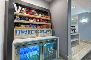 Máquina de vendas com lanches e bebidas, uma pequena geladeira com água engarrafada e uma prateleira com diversos produtos embalados.