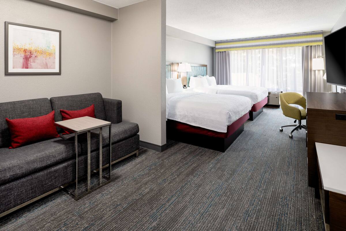 A hotel room with a grey sofa, a small table, two beds, a TV, and a desk.