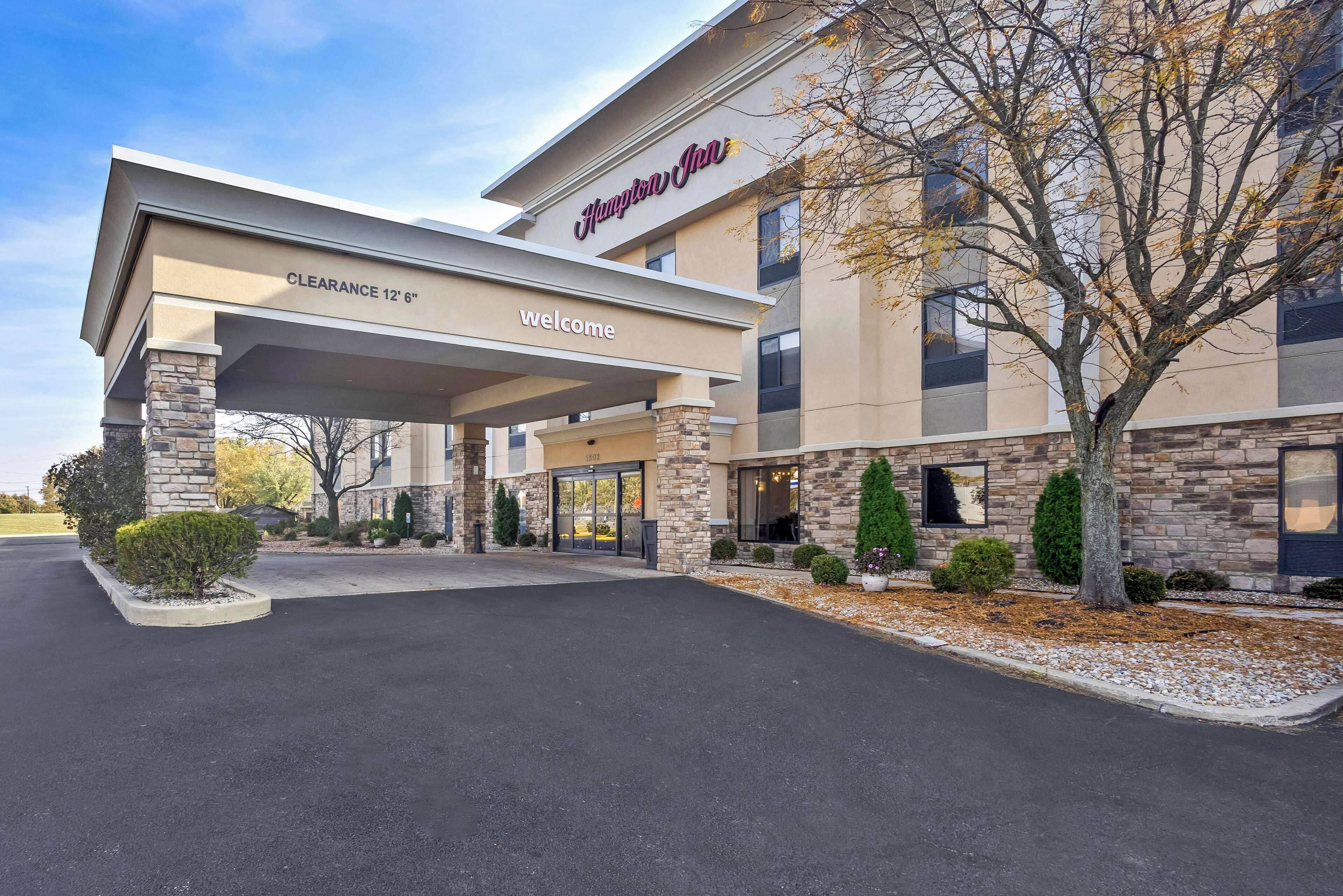 Un Hampton Inn con entrada despejada, fachada de piedra y un cartel de bienvenida.