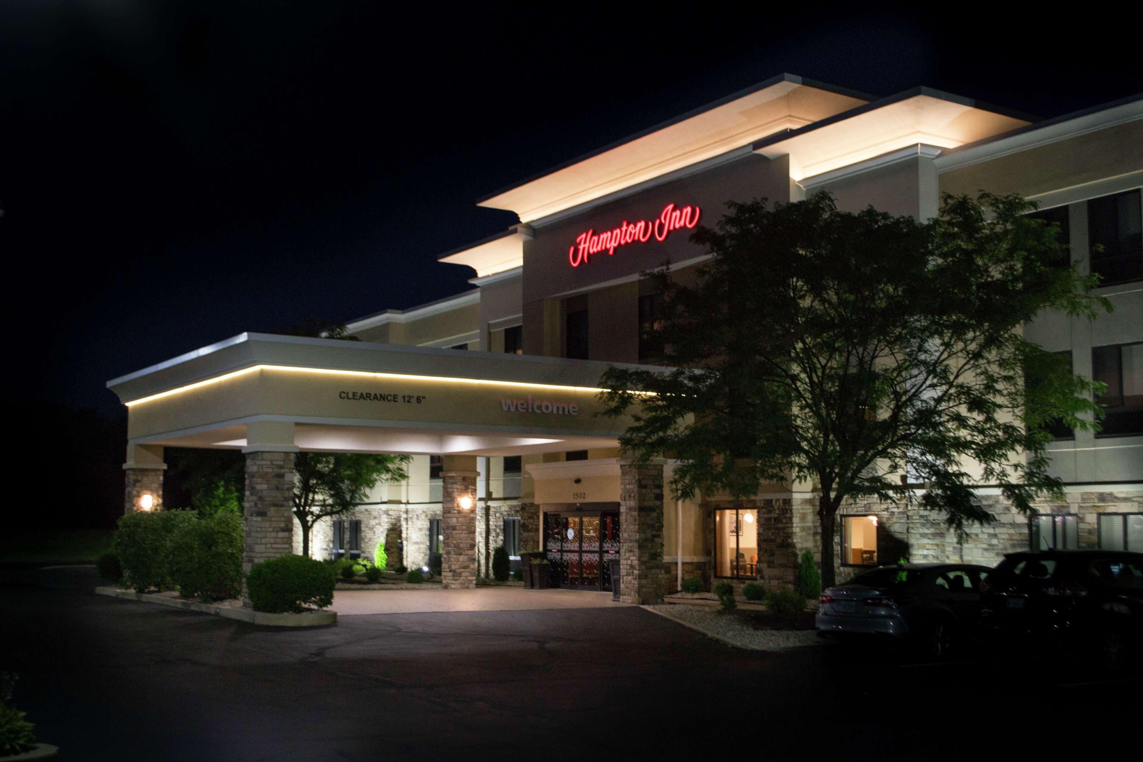 A Hampton Inn hotel entrance at night with a clear sign, a covered walkway, and a parking area.