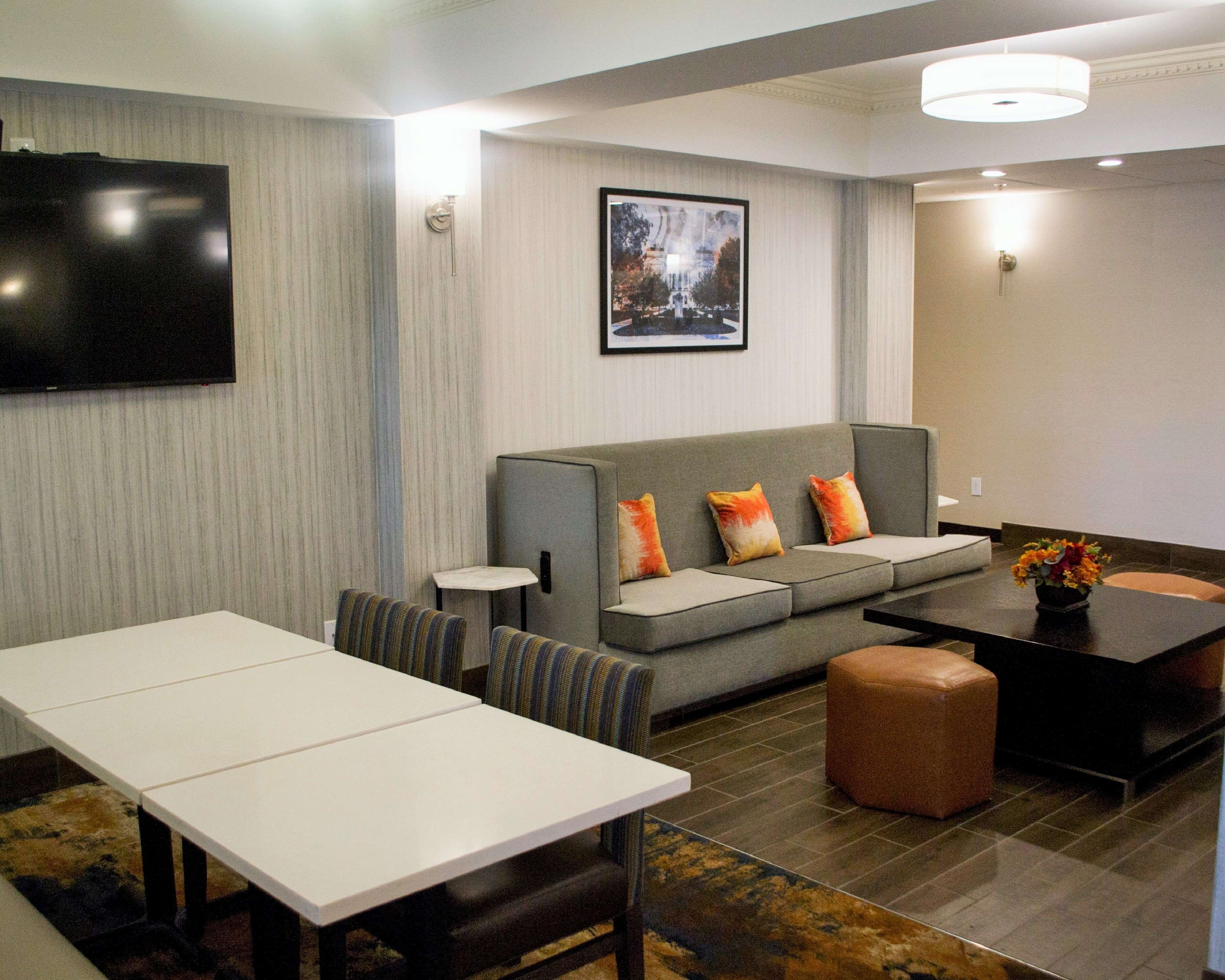 A modern hotel room with a grey sofa, striped chairs, a white table, a brown ottoman, a black coffee table, a flat-screen TV, and a framed picture on the wall.