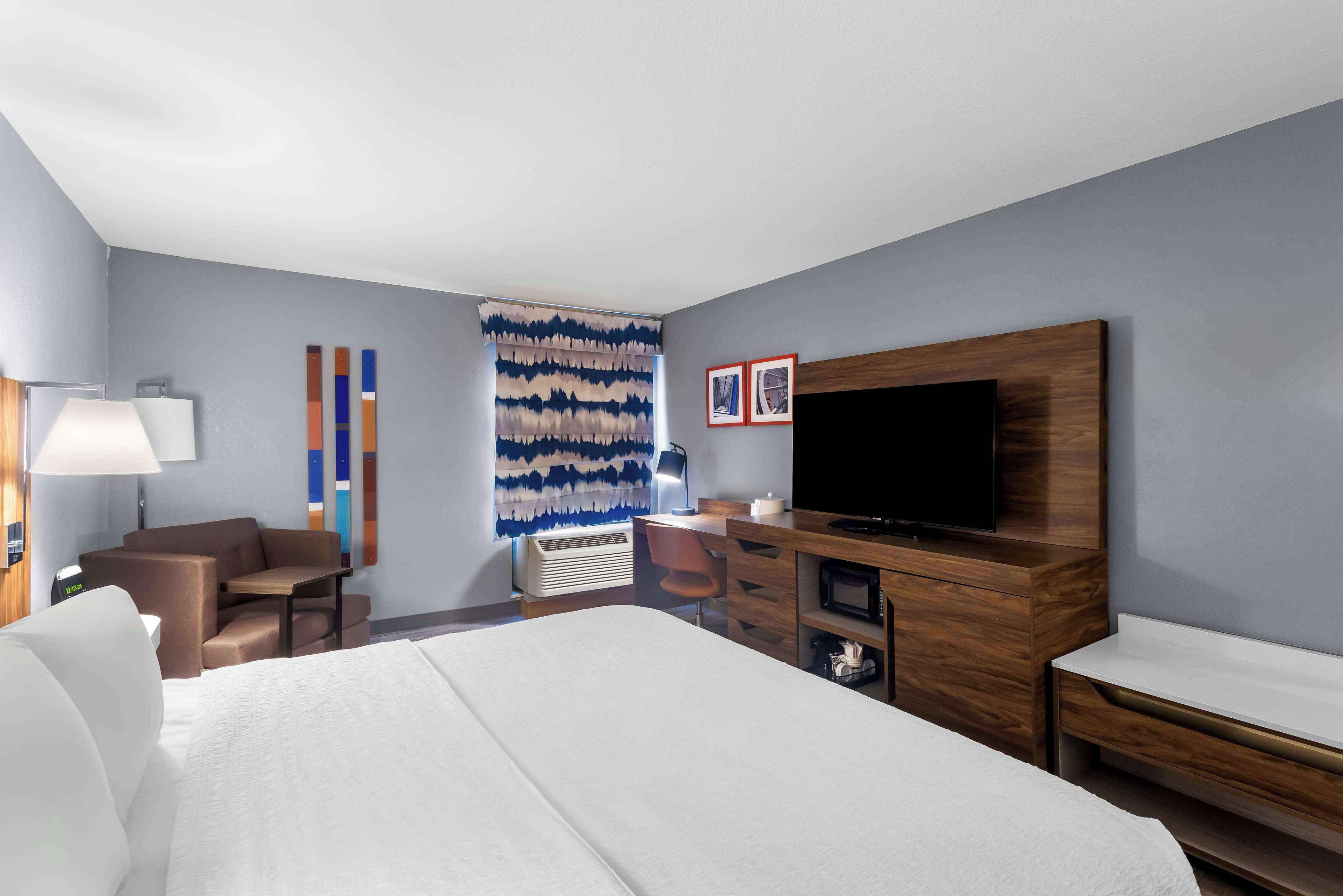 Habitación de hotel con cama, escritorio, silla, televisor y un muro con arte abstracto.