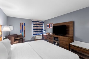 Habitación de hotel con cama, escritorio, silla, televisor y un muro con arte abstracto.