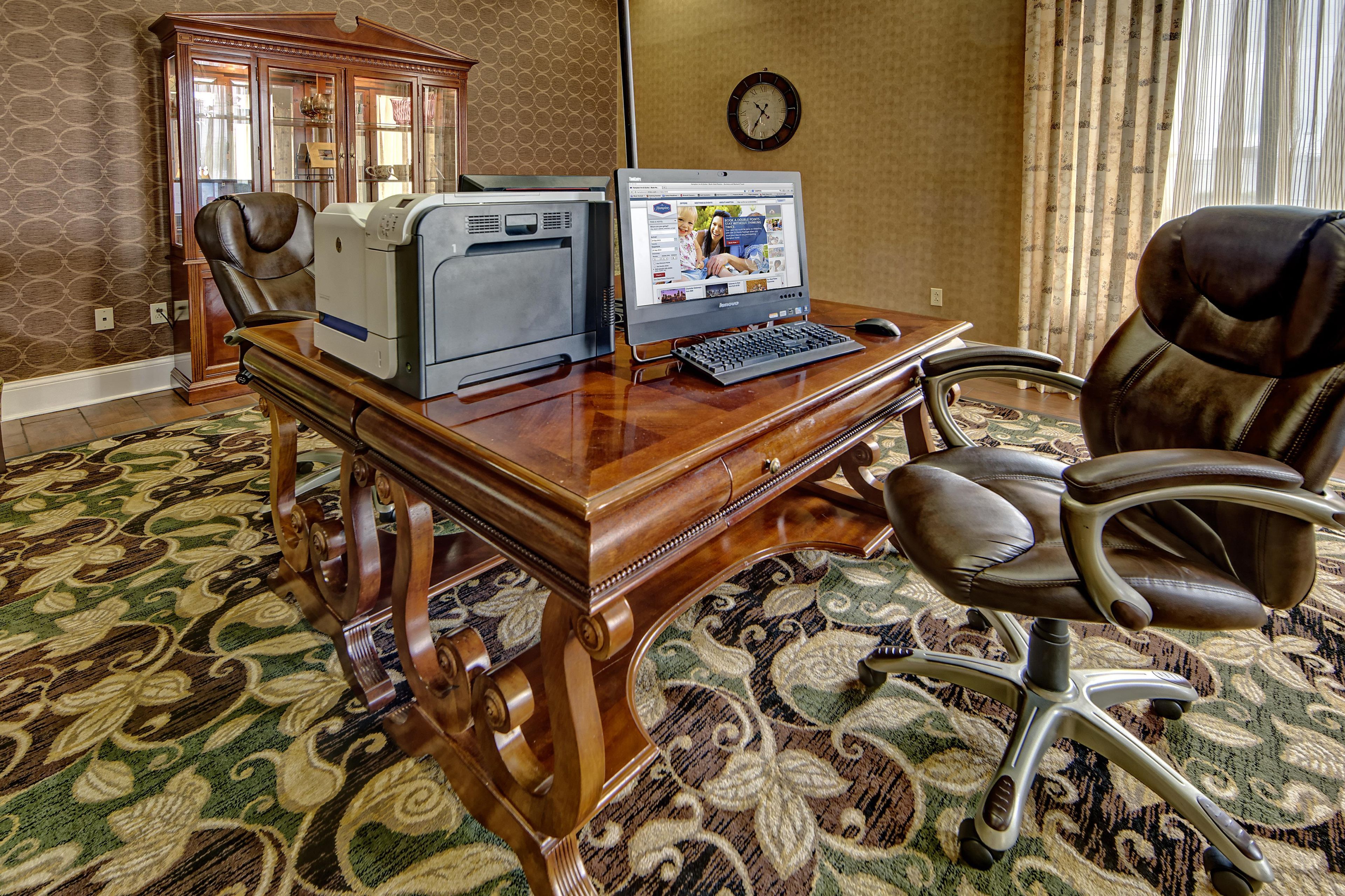 Un bureau en bois avec une imprimante, un ordinateur portable et une chaise, dans une pièce dont le sol est recouvert d’une moquette à motifs et où l’on peut voir une horloge au mur.
