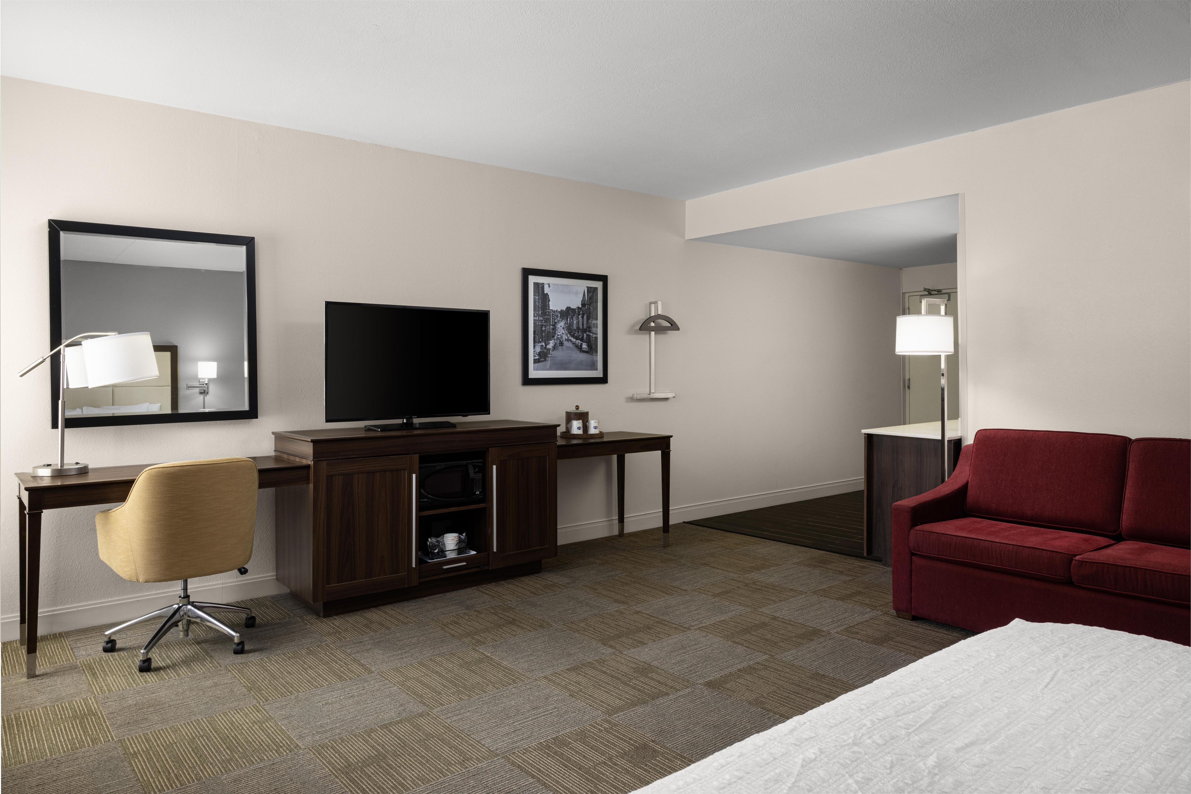 A hotel room with a bed, a desk with a TV, a chair, a red sofa, and a mirror.