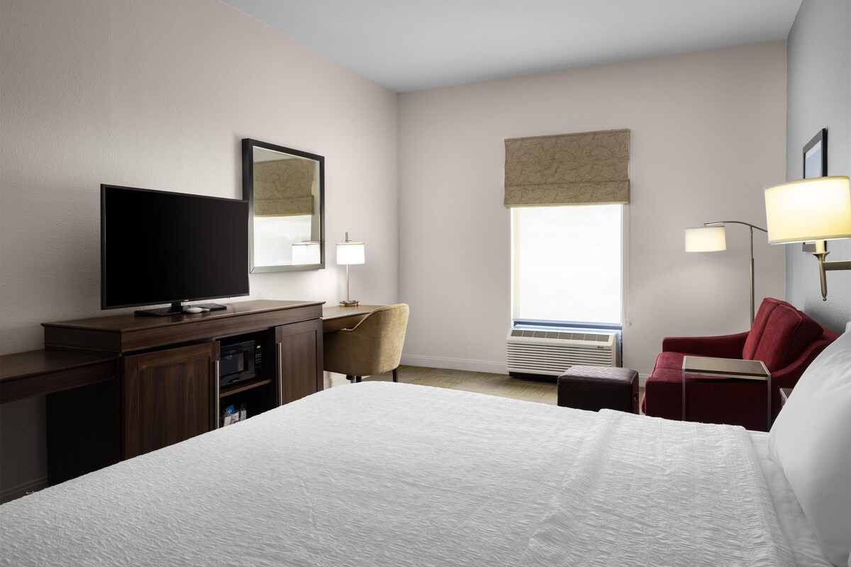 Una habitación de hotel con cama, escritorio, televisión, silla, lámpara y ventana.