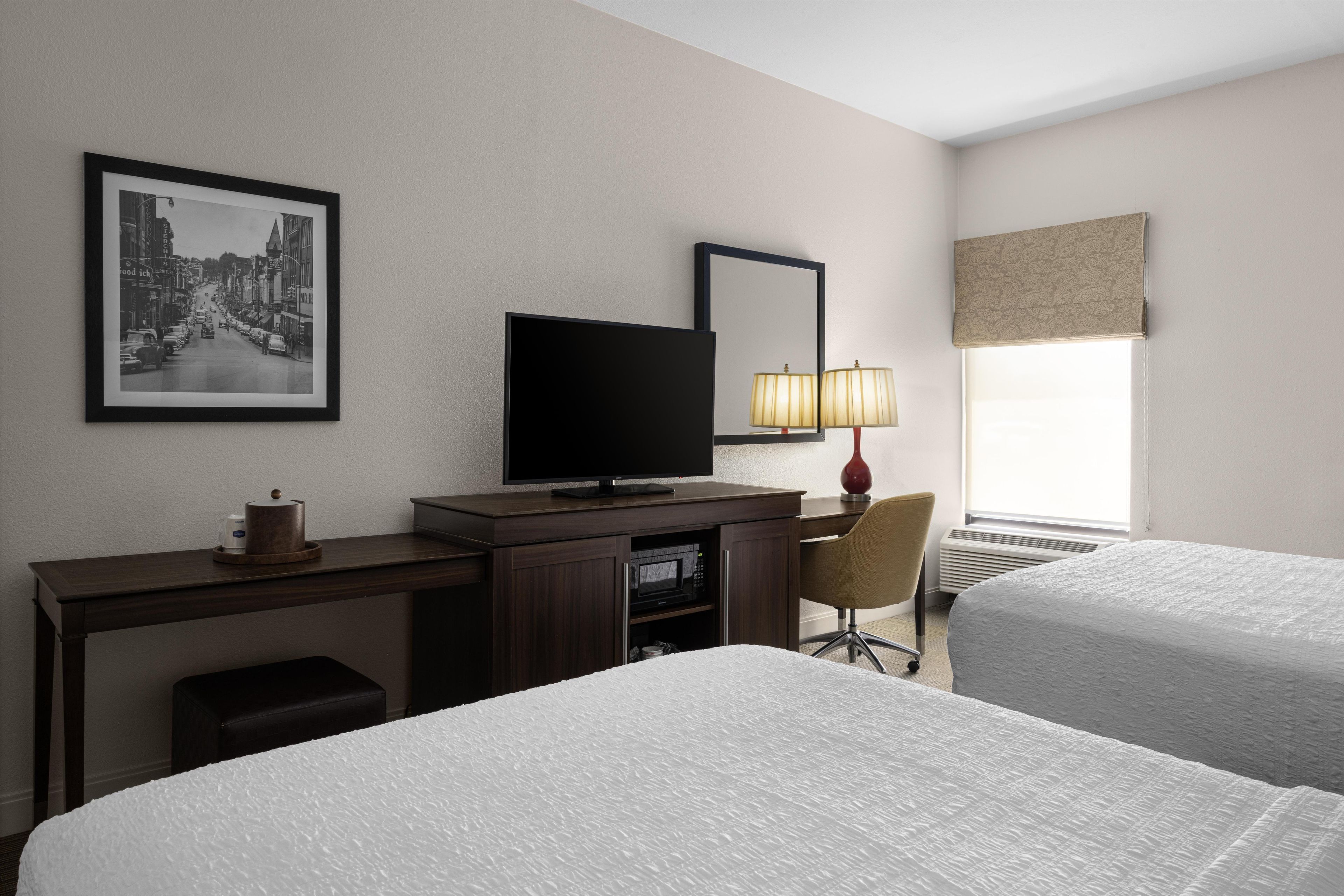 A hotel room with two beds, a desk with a television, a chair, a lamp, and a framed picture on the wall.