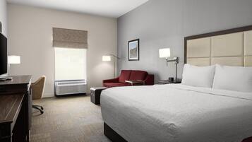 A hotel room with a large bed, a desk, a chair, a red armchair, a flat-screen TV, and a window with a view.
