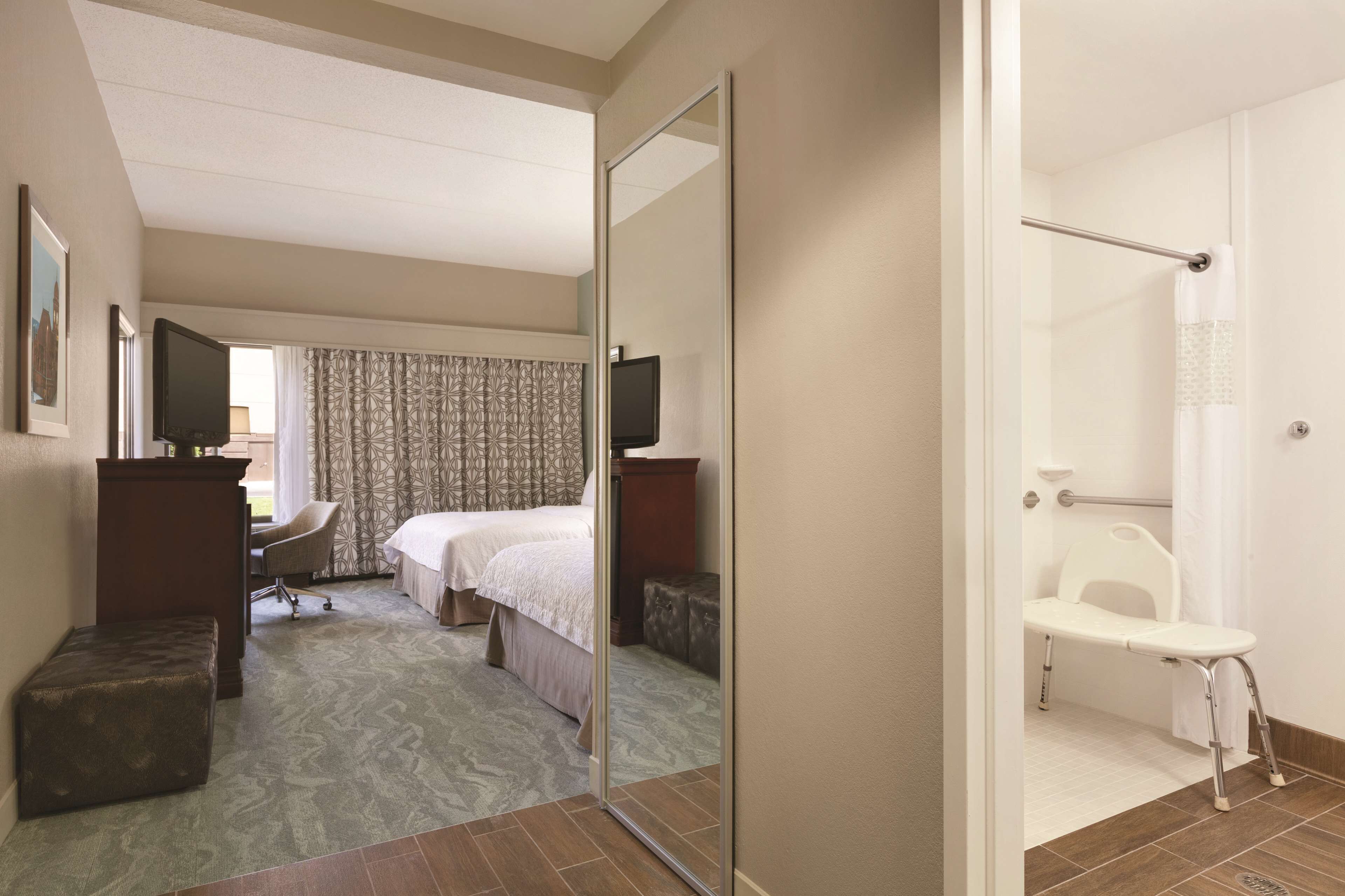 Habitación de hotel con cama, escritorio, silla, televisor y zona de ducha con banco.