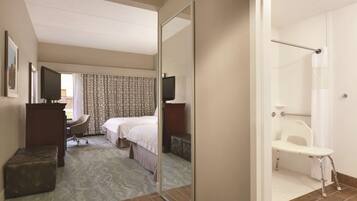 Habitación de hotel con cama, escritorio, silla, televisor y zona de ducha con banco.