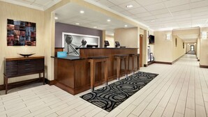 Een hotel receptie met een houten balie, barstoelen, een digitaal scherm en een receptiebalie.