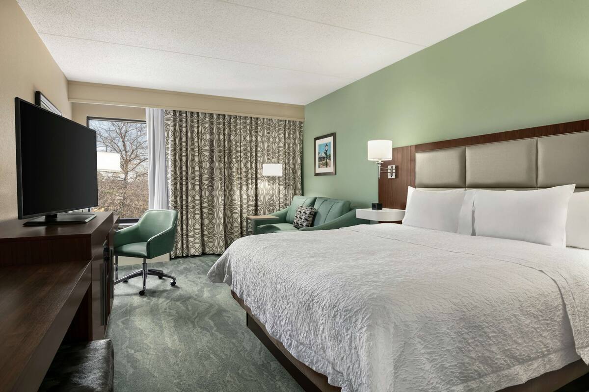 Une chambre d’hôtel avec un lit, un bureau avec une télévision, un fauteuil vert, une fenêtre avec des rideaux et une tête de lit agrémentée de lampes.