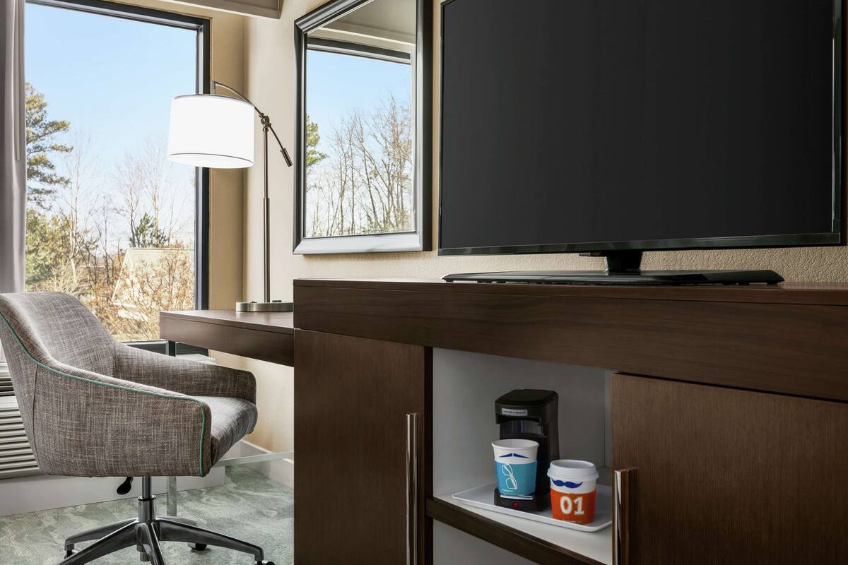 Une chambre d’hôtel dotée d’une grande fenêtre, d’un bureau avec un téléviseur à  écran plat, d’une chaise, d’une lampe et d’une machine à café.