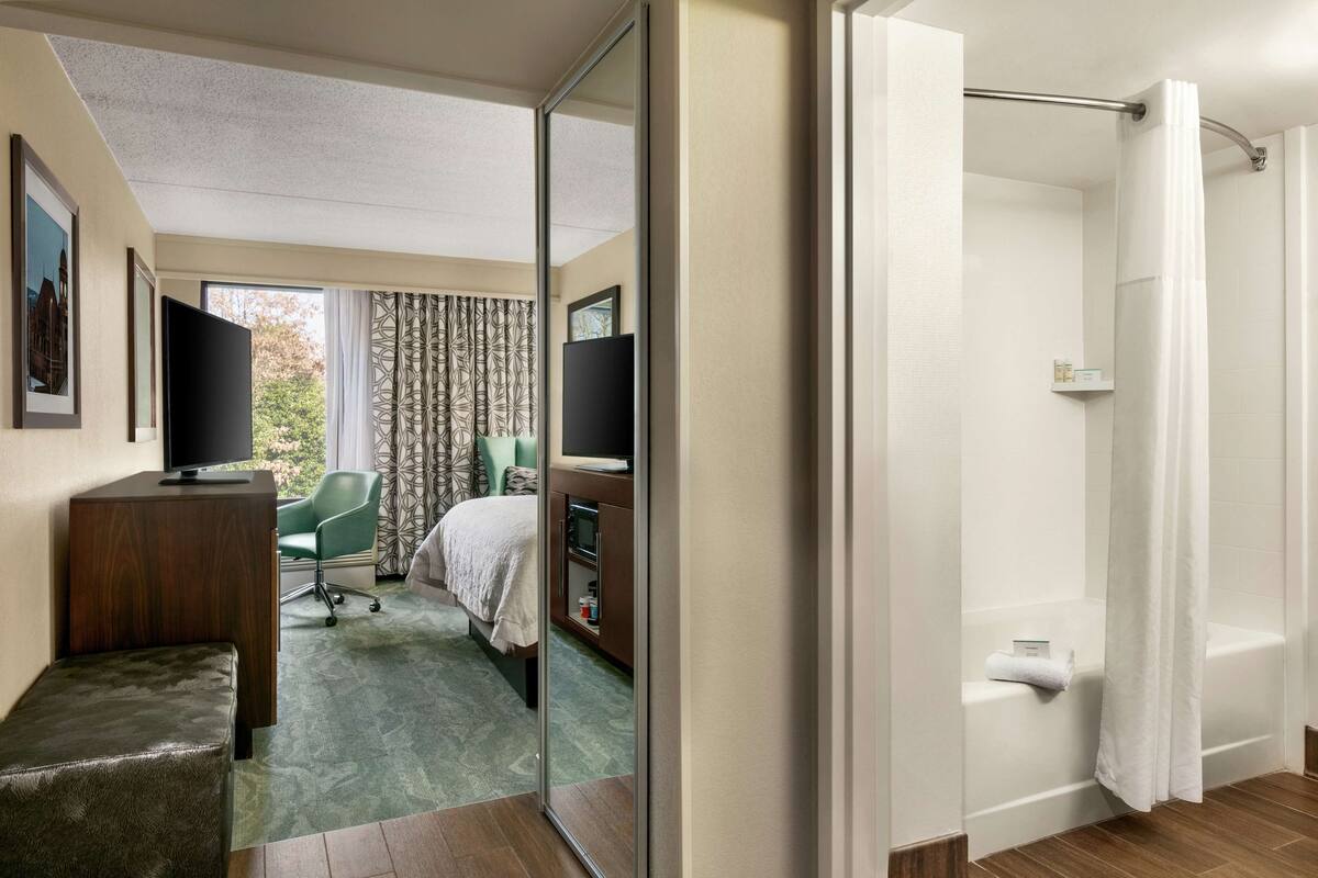 Une chambre d’hôtel comprenant un lit, un bureau avec une télévision, une chaise, un miroir et un espace douche.