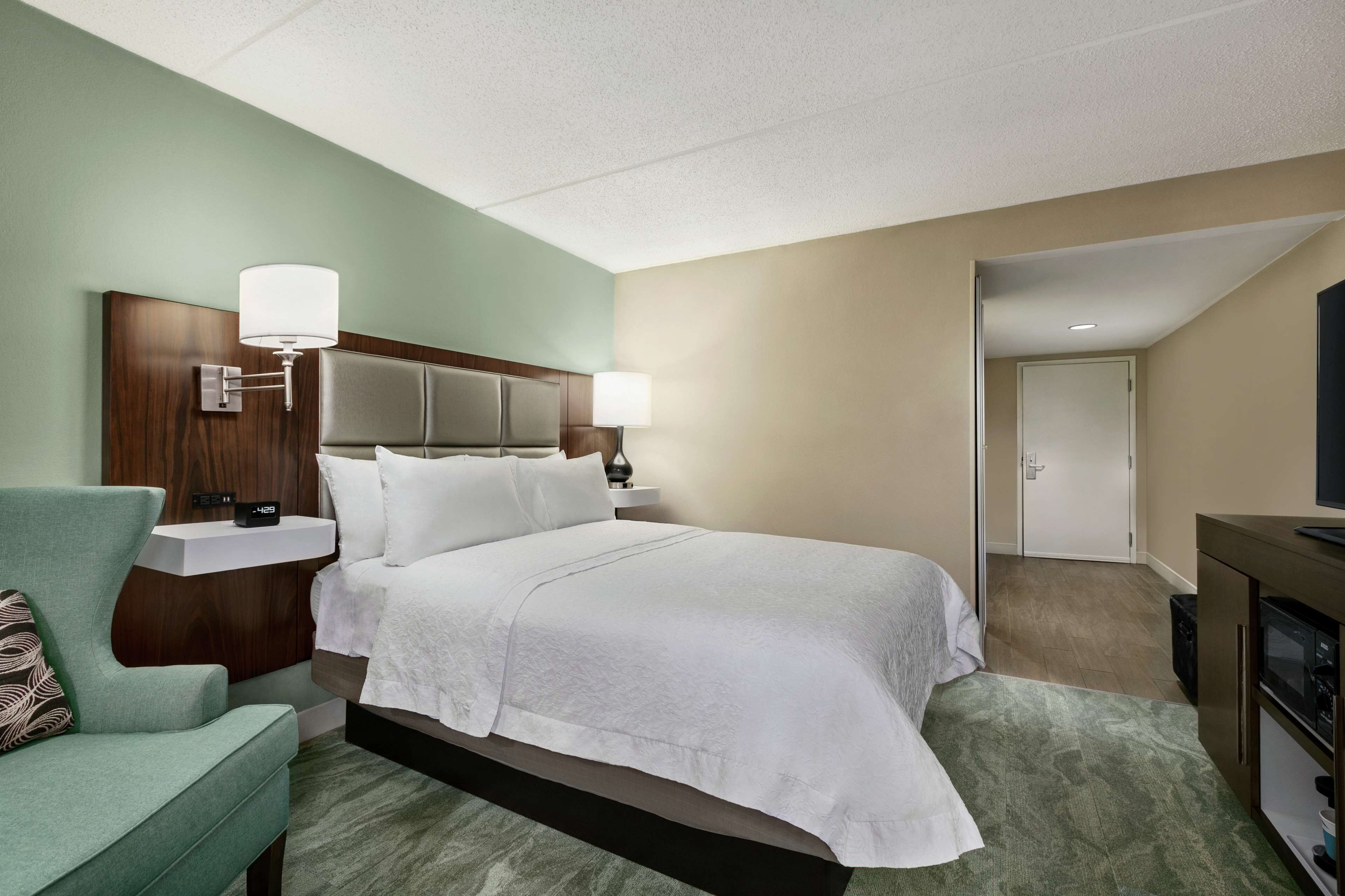 Une chambre d’hôtel avec un grand lit, une table de chevet avec une lampe, une télévision et un coin salon avec un fauteuil.