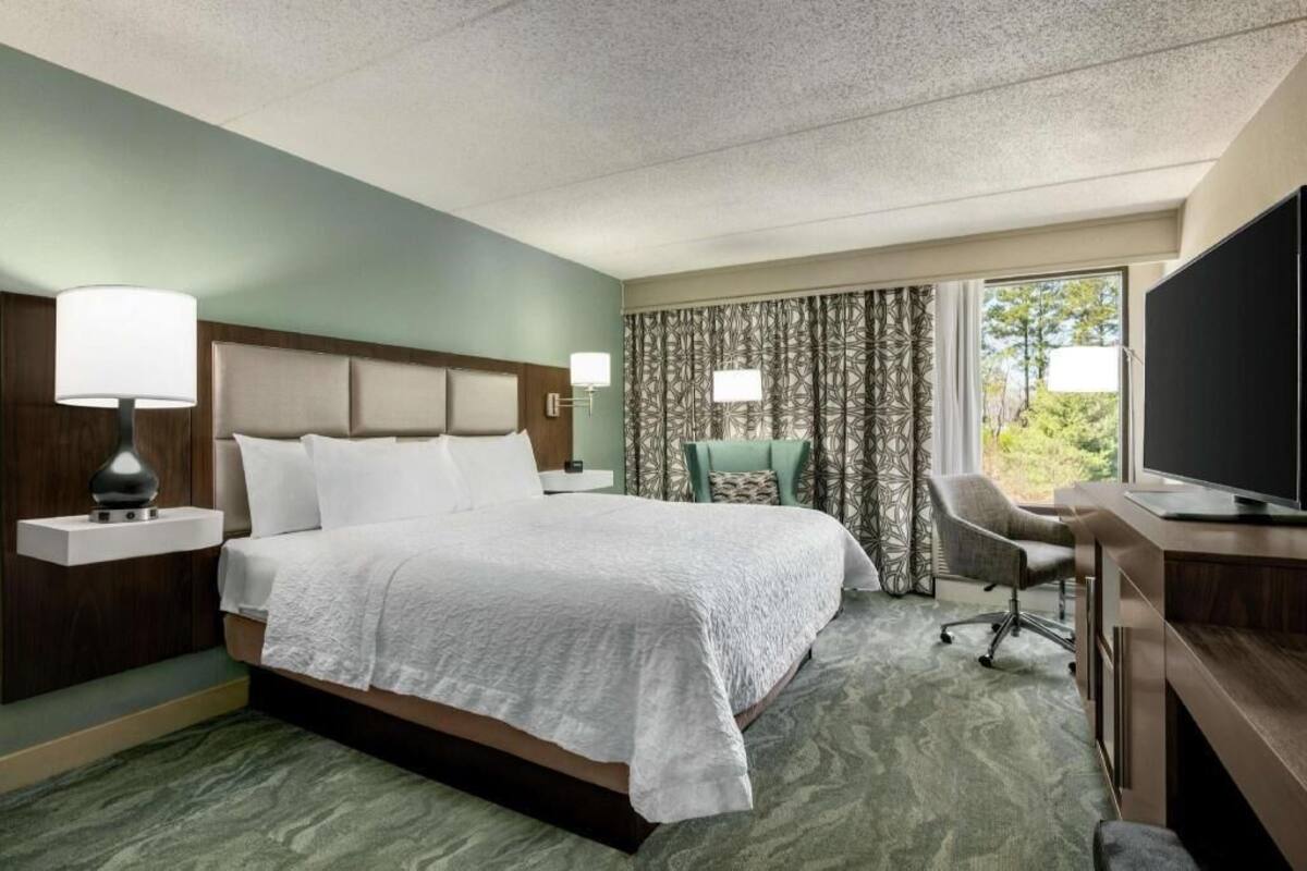 Une chambre d’hôtel avec un lit, un bureau, une chaise, une télévision et une fenêtre avec des rideaux.