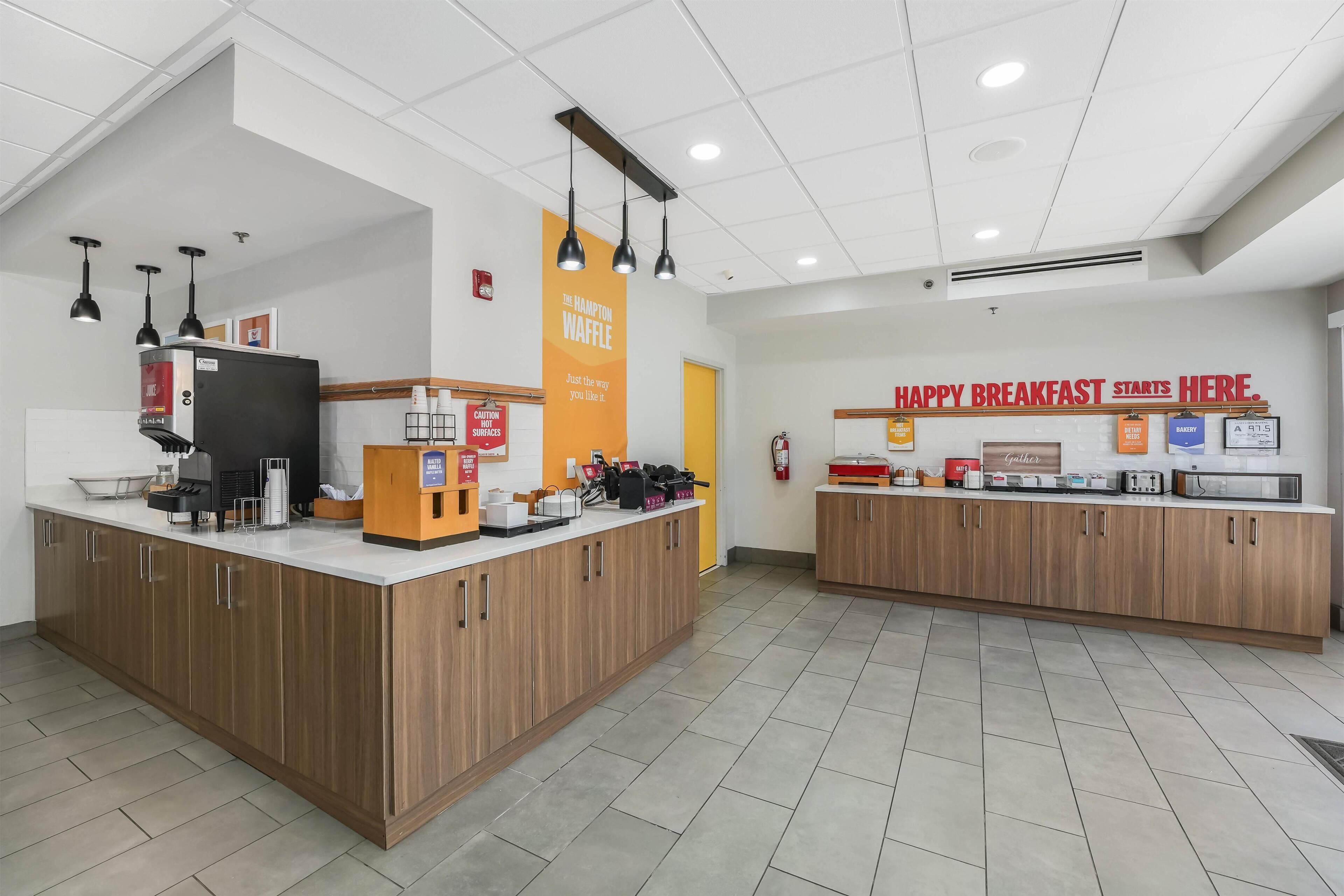 Uma área de café da manhã com máquina de café, estação de waffles e um letreiro que diz 'CAFÉ DA MANHÃ FELIZ COMEÇA AQUI'.
