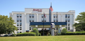 A Hampton Inn by Hilton hotel with a white facade, multiple windows, and an American flag.