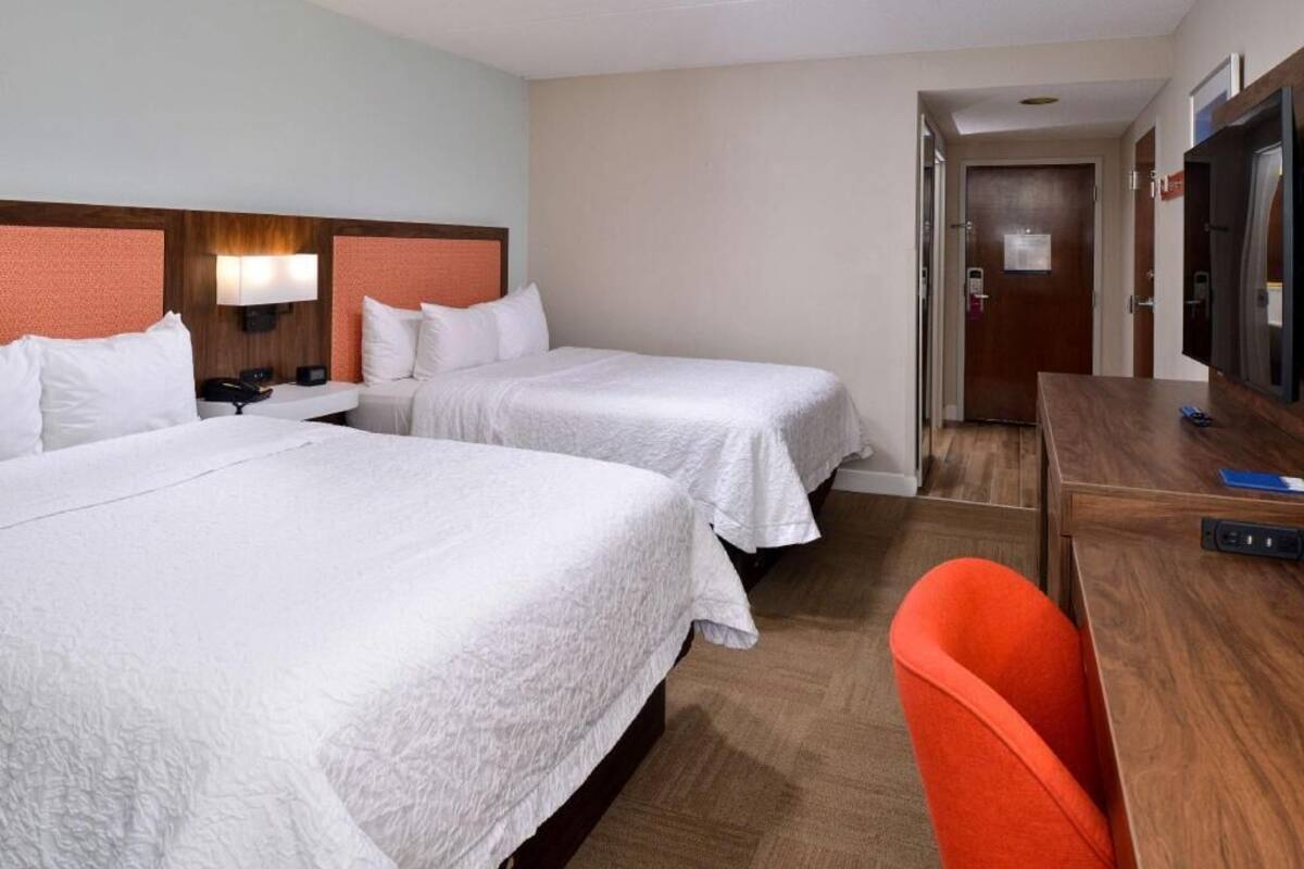 Quarto de hotel com duas camas, uma escrivaninha, uma cadeira e uma porta.