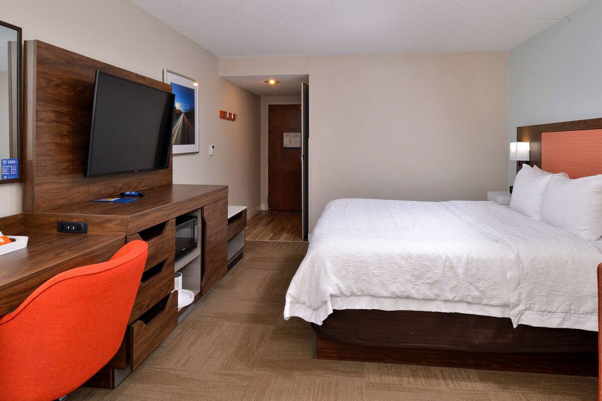Quarto de hotel com cama, escrivaninha com televisão, cadeira e porta que dá para outro cômodo.