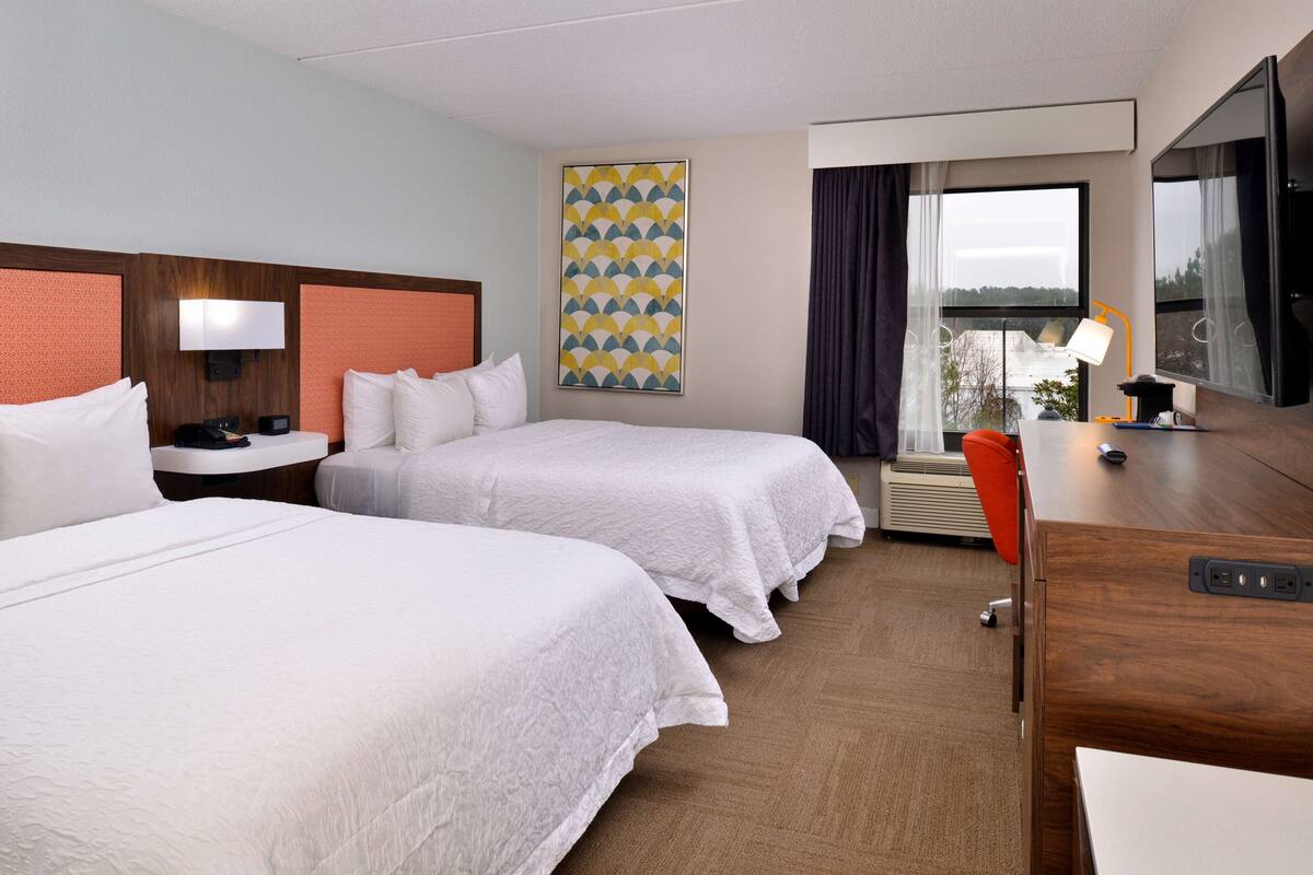 Quarto de hotel com duas camas, uma escrivaninha, uma cadeira, uma televisão e janela com cortinas.