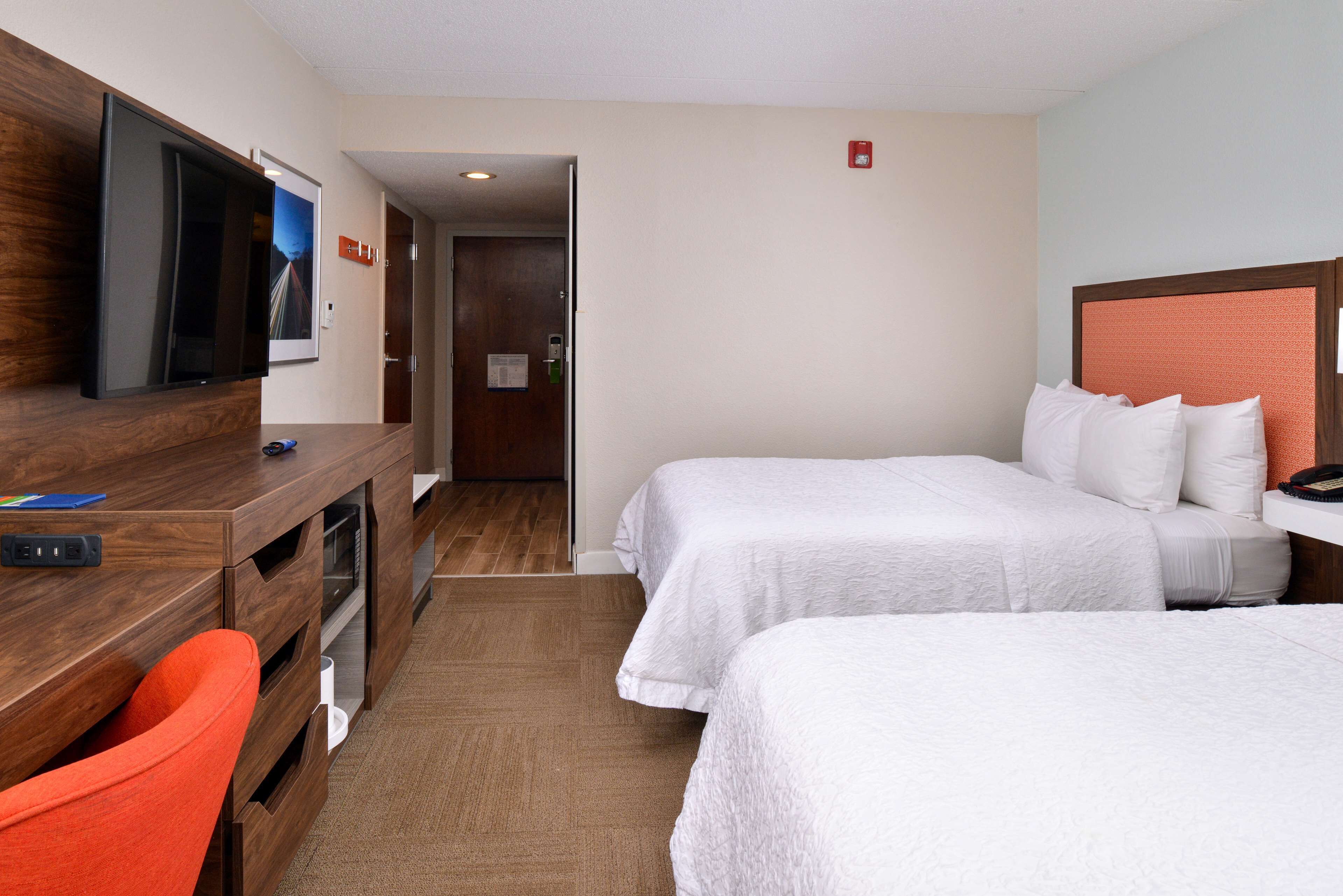 A hotel room with two beds, a desk with a flat-screen TV, a red chair, and a wooden dresser.