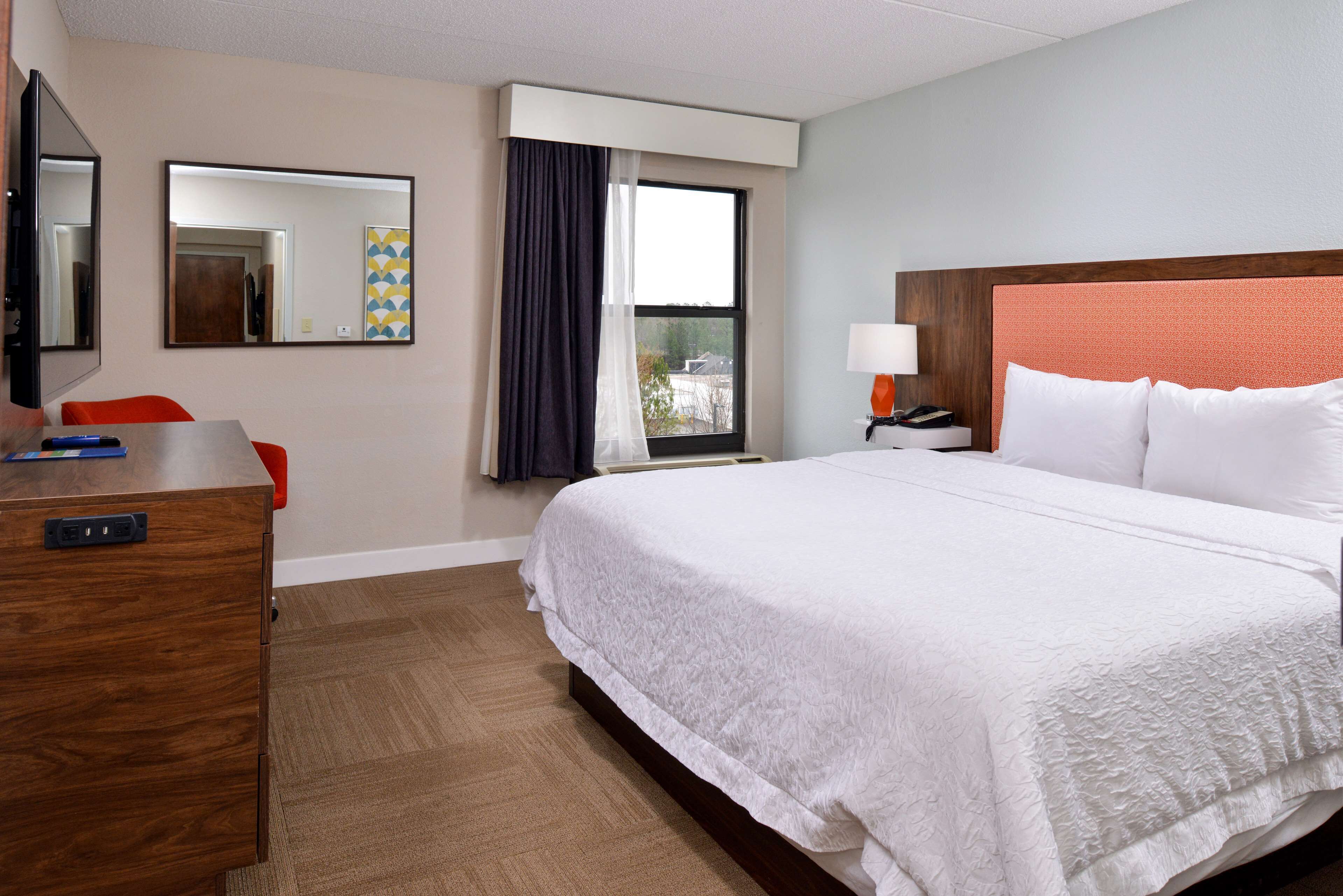 Quarto de hotel com uma cama grande, uma mesa com cadeira, uma televisão, uma janela com cortinas e uma mesa de cabeceira com luminária.