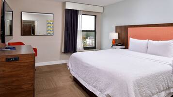 Quarto de hotel com uma cama grande, uma mesa com cadeira, uma televisão, uma janela com cortinas e uma mesa de cabeceira com luminária.