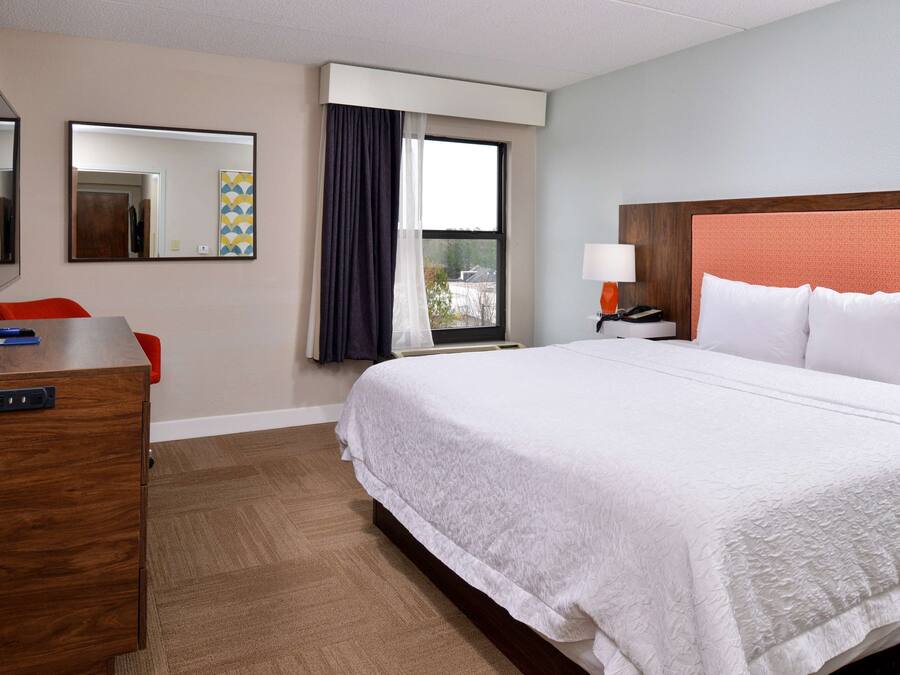Quarto de hotel com uma cama grande, uma mesa com cadeira, uma televisão, uma janela com cortinas e uma mesa de cabeceira com luminária.
