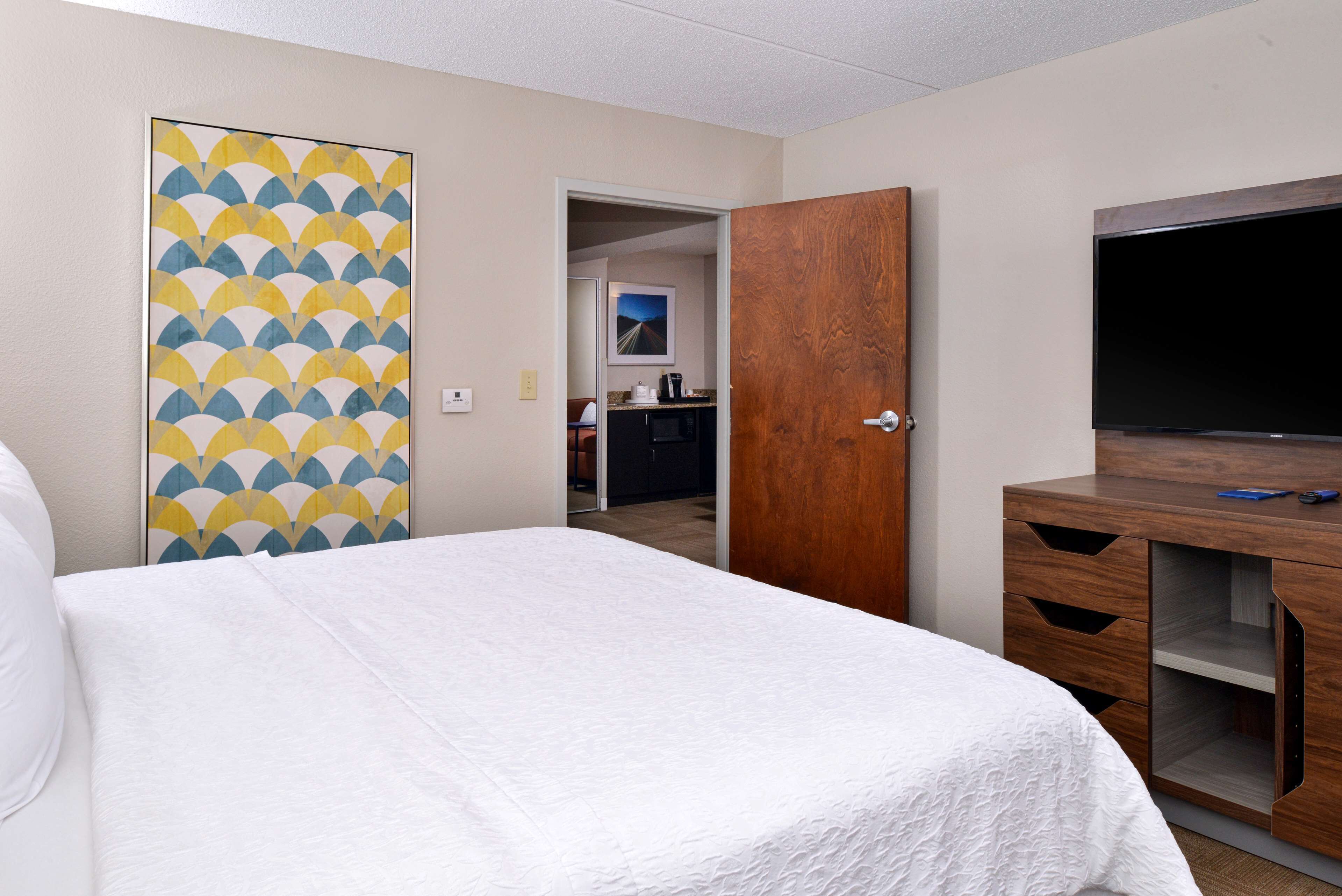 A hotel room with a bed, a wooden wardrobe, a television, and a kitchen area visible through an open door.