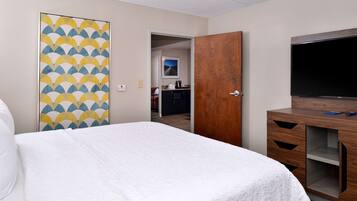 A hotel room with a bed, a wooden wardrobe, a television, and a kitchen area visible through an open door.