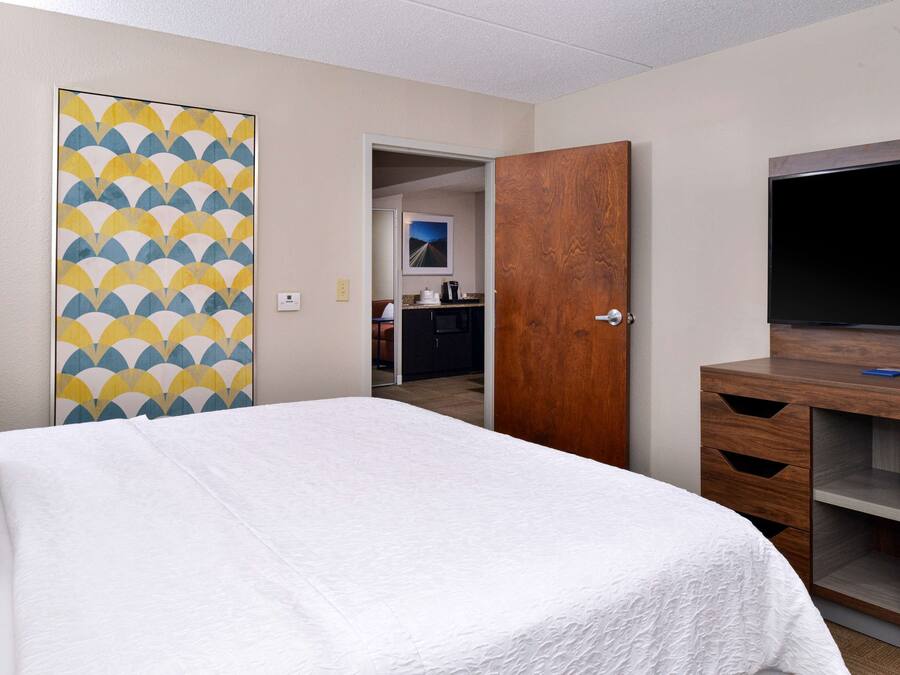 A hotel room with a bed, a wooden wardrobe, a television, and a kitchen area visible through an open door.