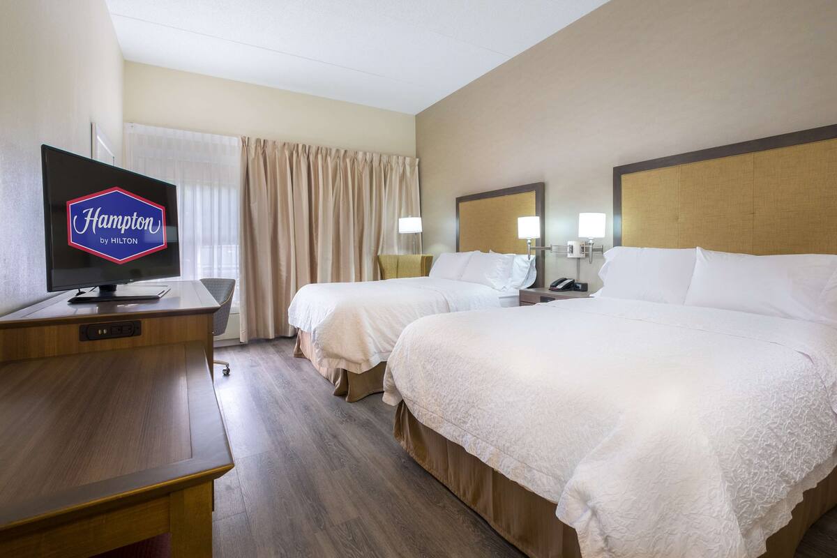 A hotel room with two beds, a desk with a television displaying 'Hampton by Hilton', and a window with curtains.