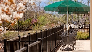 An outdoor patio with a row of tables and chairs, a green umbrella, and a wooden fence.