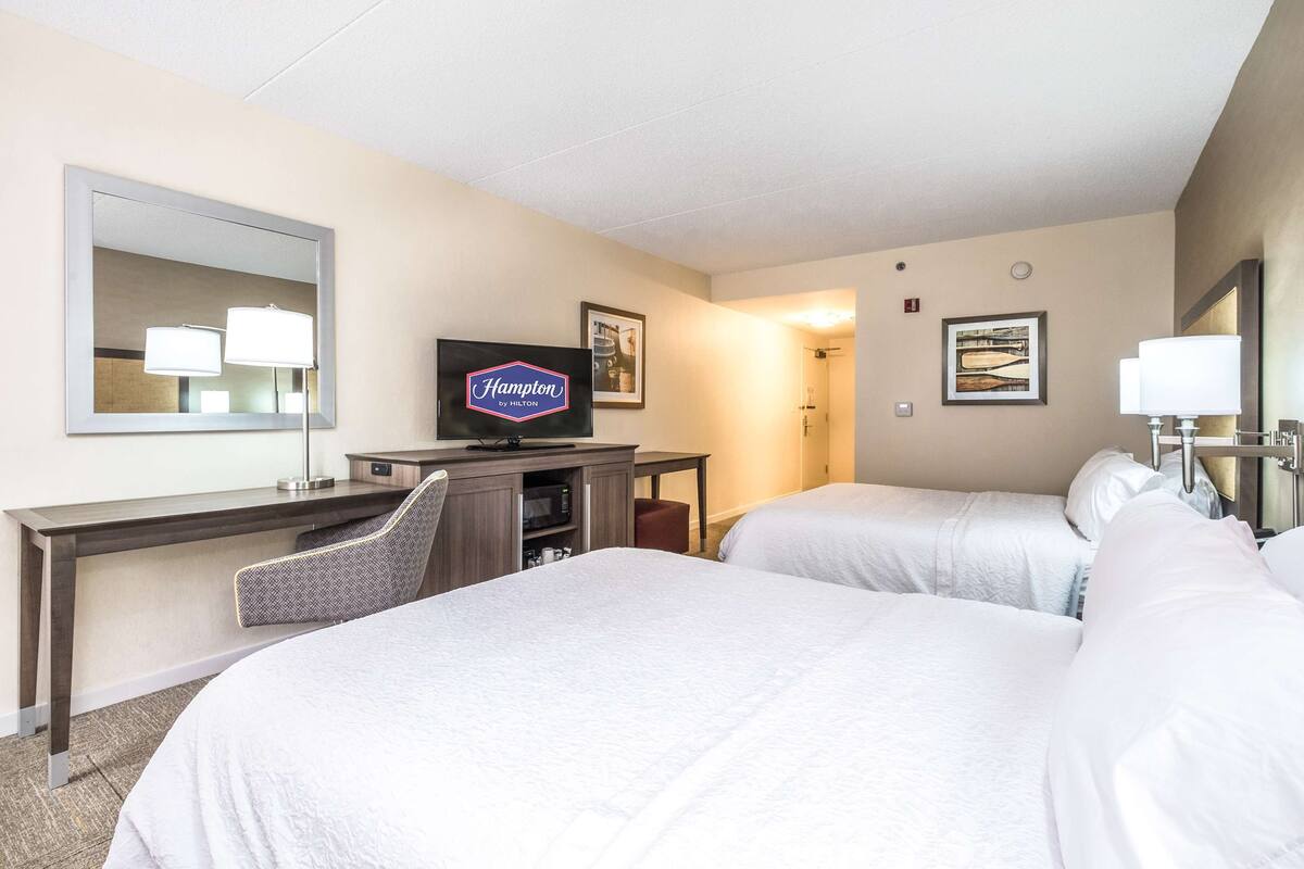 A hotel room with two beds, a desk with a television displaying 'Hampton by Hilton', a mirror, and framed artwork on the walls.