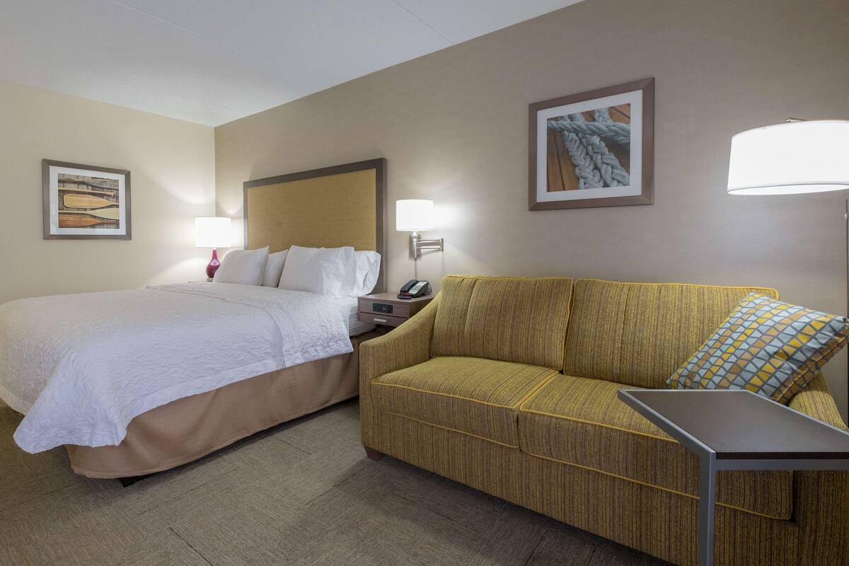 A hotel room with a bed, a sofa, a nightstand with a lamp, and two framed pictures on the wall.