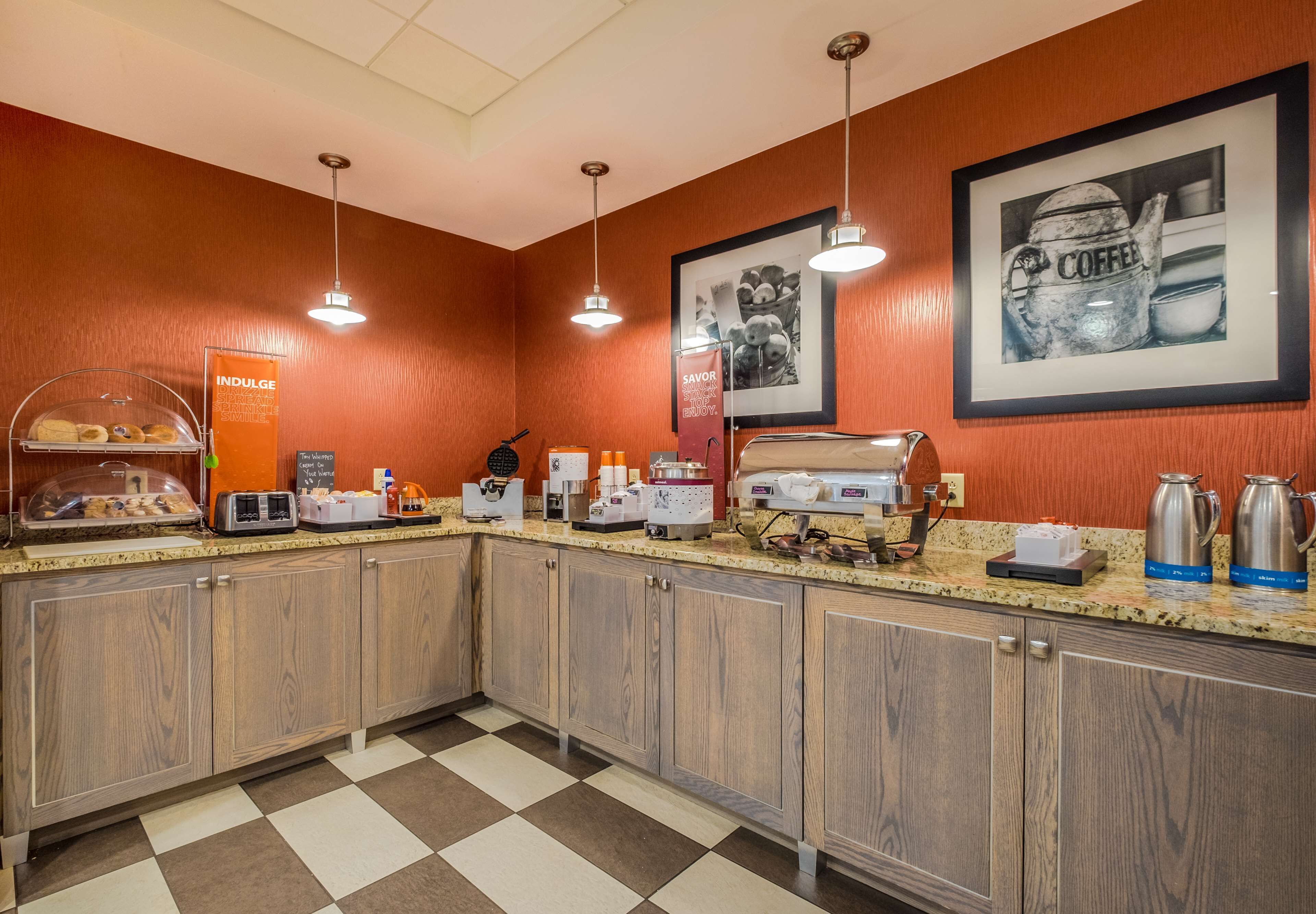 Une cuisine d’hôtel avec des armoires en bois, un sol à carreaux, un coin café et une œuvre d’art encadrée sur le thème du café accrochée au mur.