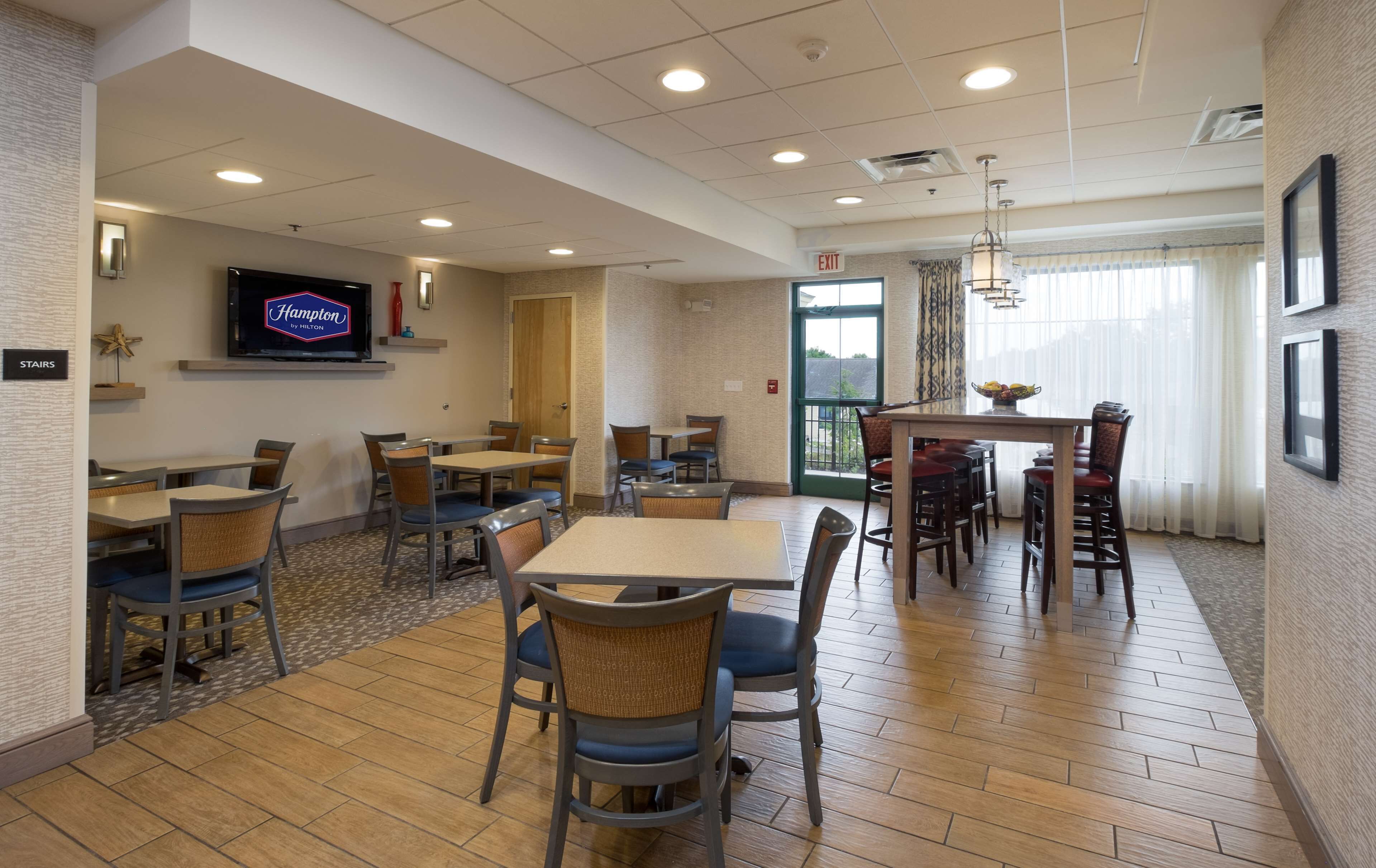 A hotel dining area with multiple tables and chairs, a flat-screen TV mounted on the wall, and a view of the outdoors through a large window.