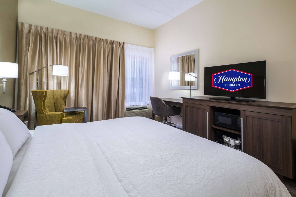 A hotel room with a large bed, a desk, a chair, a television, and a minibar.