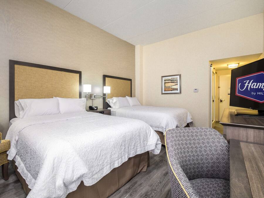 Una habitación de hotel con dos camas, una silla, un escritorio, un televisor y un cuadro en la pared.