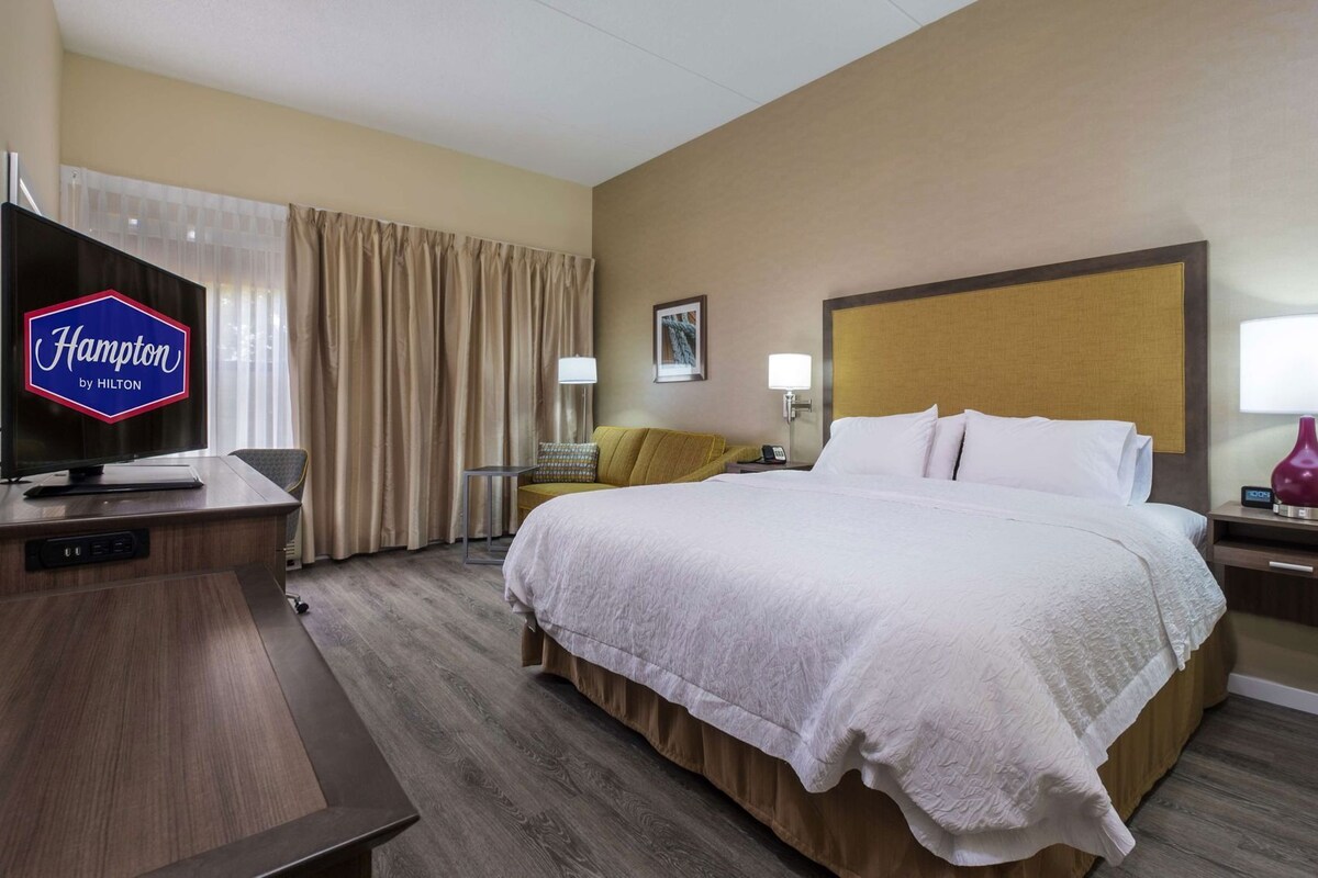 A hotel room with a bed, a desk with a television, a nightstand with a lamp, and a wall-mounted picture.