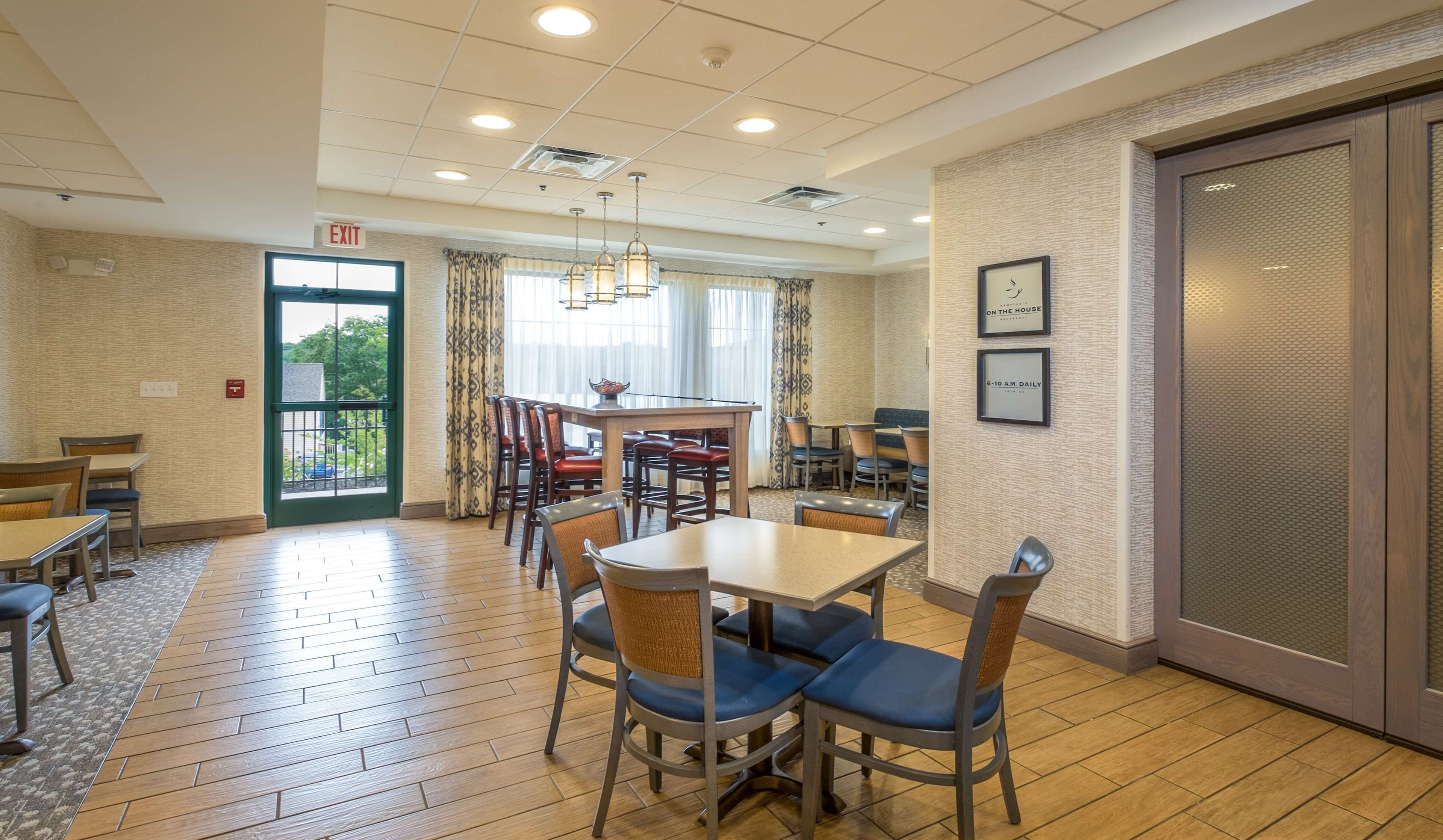 A hotel dining area with a central table, chairs, and a bar. There is an exit sign above the door, and a framed sign on the wall.