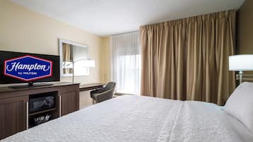Una camera d'albergo con un letto, una scrivania, una televisione che mostra 'Hampton by Hilton', una lampada e una finestra con tende.