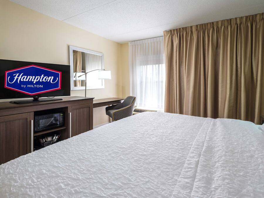 Una camera d'albergo con un letto, una scrivania, una televisione che mostra 'Hampton by Hilton', una lampada e una finestra con tende.